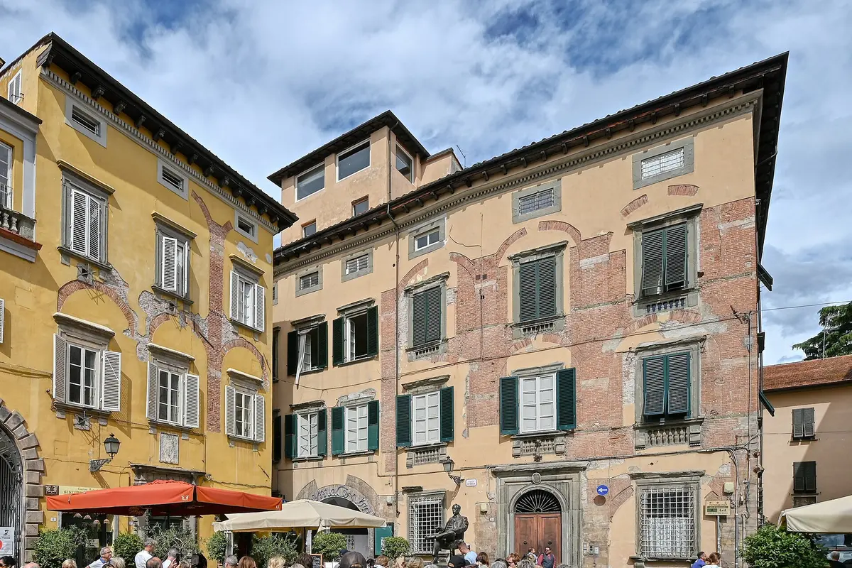 Two tall old palazzi with ochre and pink plaster walls and green shutters around a small Lucca square, with café umbrellas at street level and a blue sky above