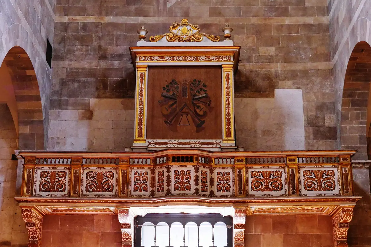 A painted and gilded wooden organ loft high on the wall of a Romanesque church, with a carved balcony front and a small barred window beneath it.