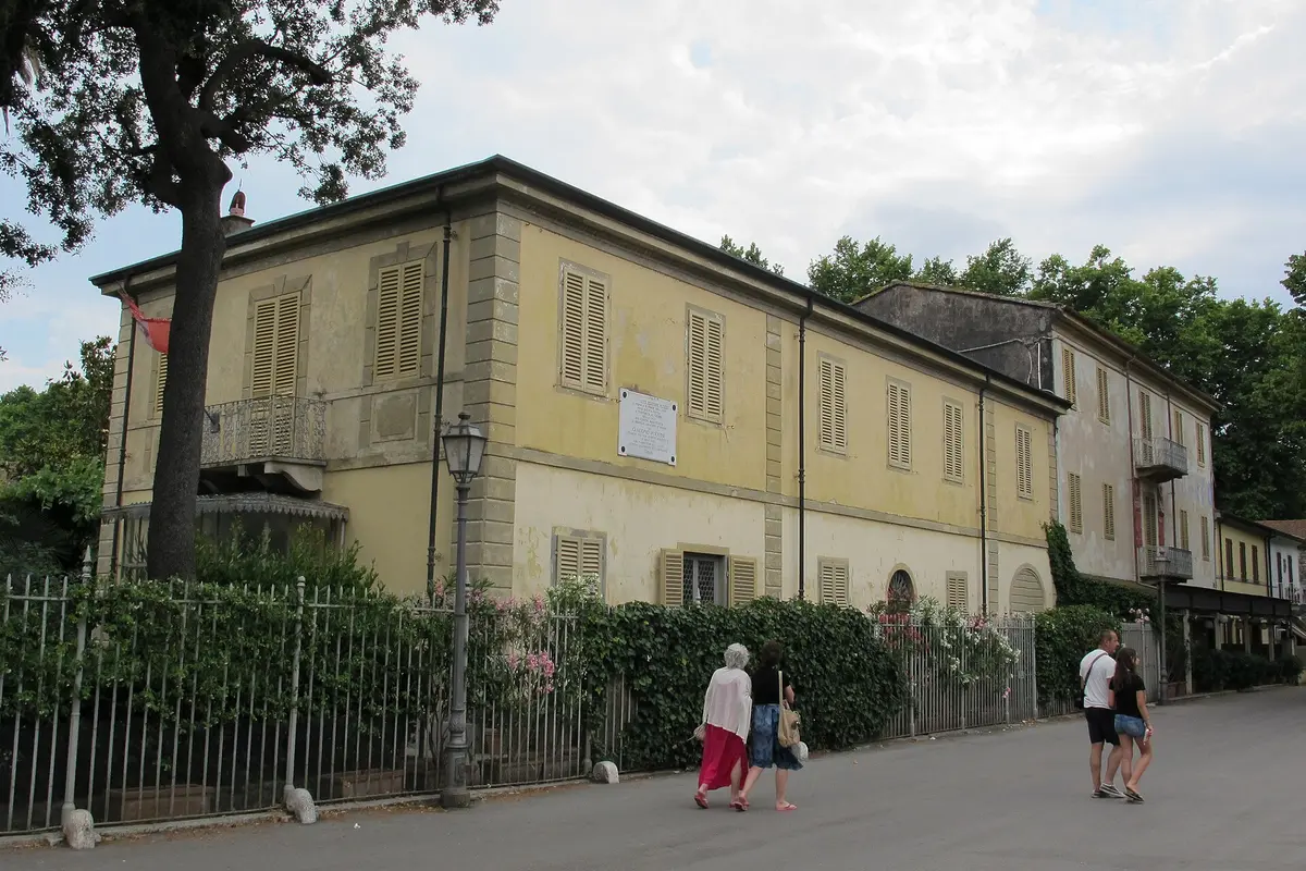 A long pale-yellow villa with green shutters behind a low iron fence, with tall trees beside it and two people walking past on the pavement