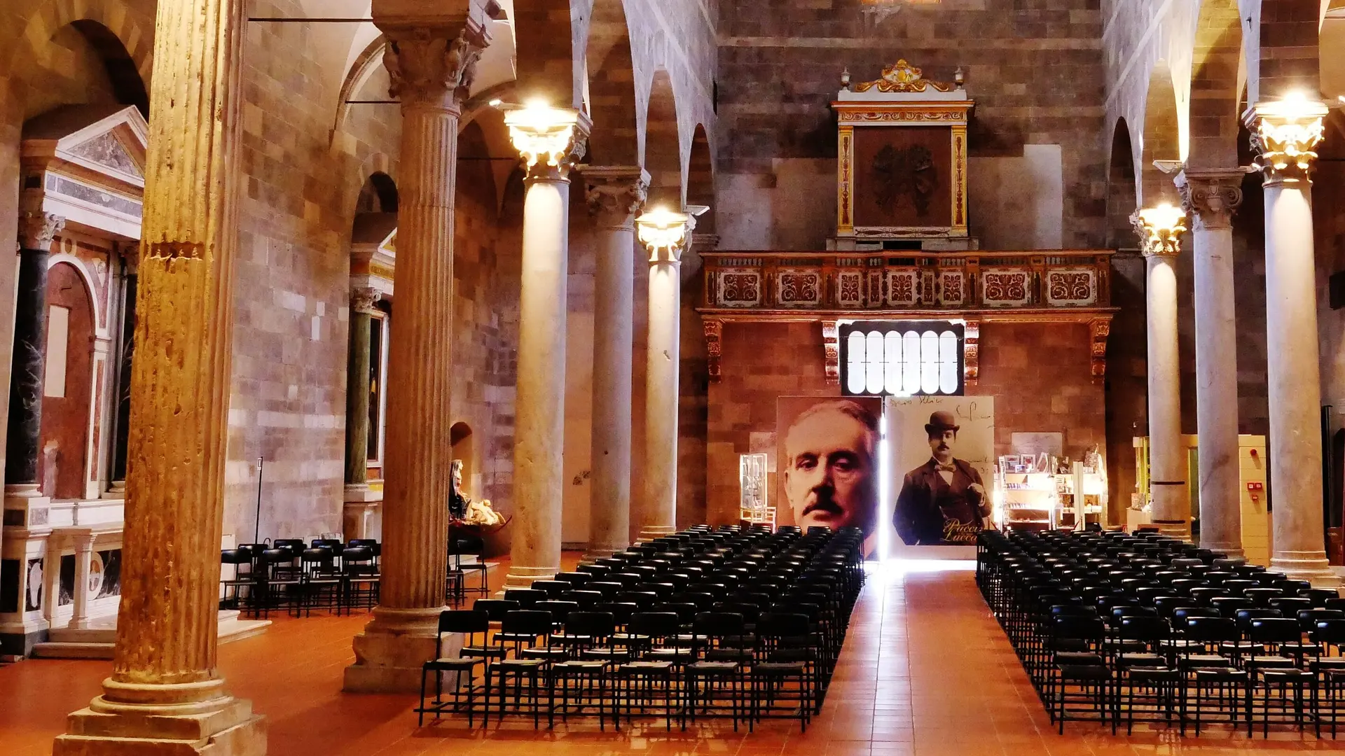 The nave of a Romanesque church in Lucca with rows of black chairs facing a gilded organ loft, beneath which two large portraits of Giacomo Puccini stand on boards, lit by warm lamps on the columns.